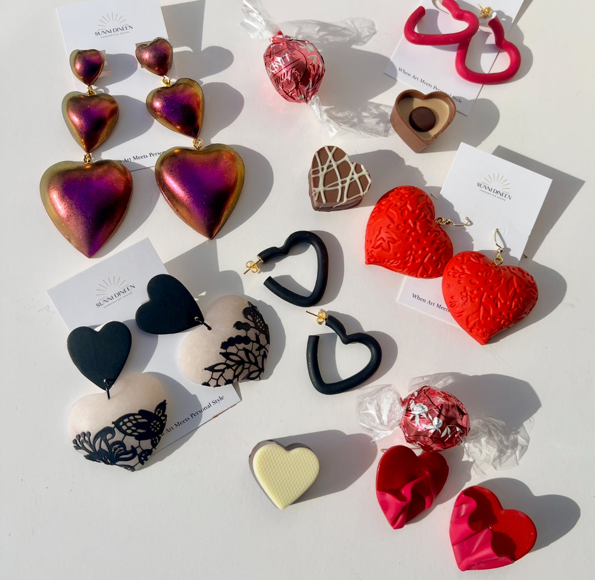 The Valentine's Day Collection featuring heart-shaped statement earrings handmade by Sunni Dineen,  along with heart-shaped chocolate candies on a white surface.