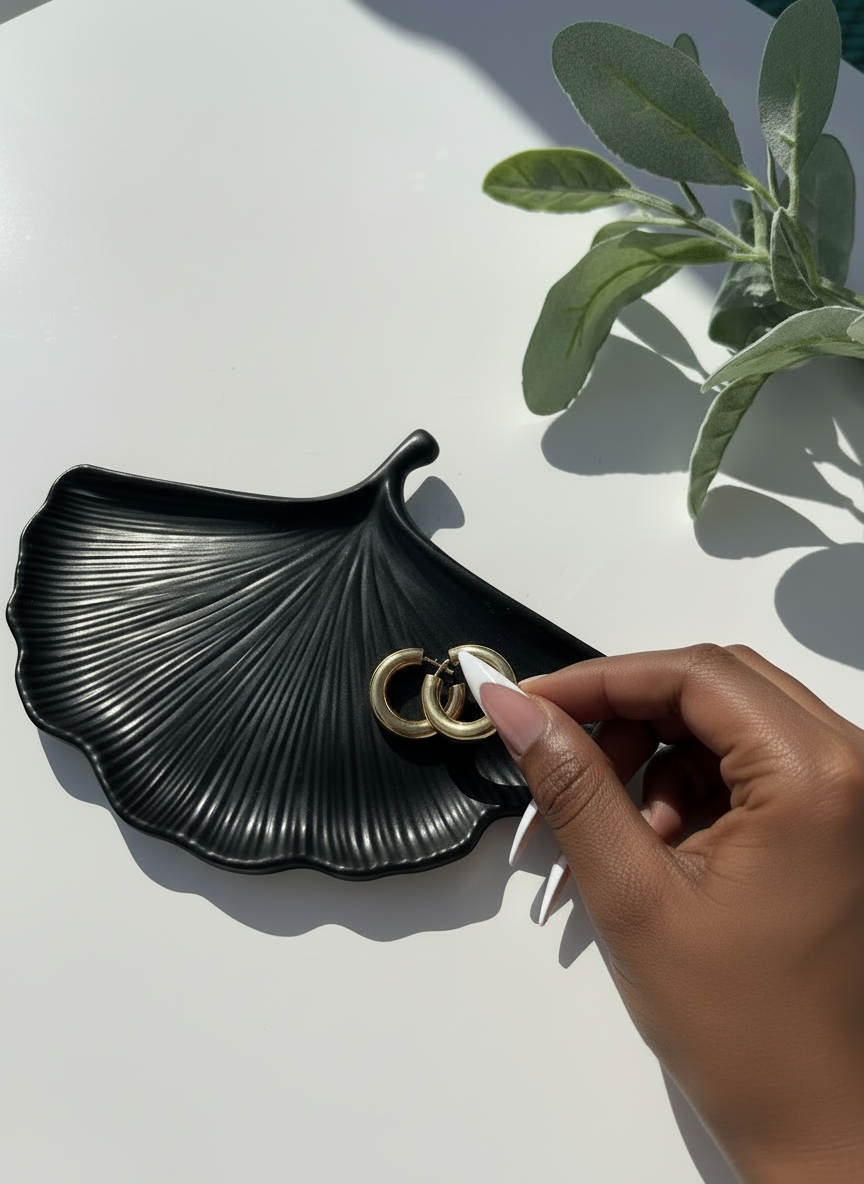 Black Ginkgo Trinket Dish with a hand placing gold hoop earrings on a light surface with greenery.