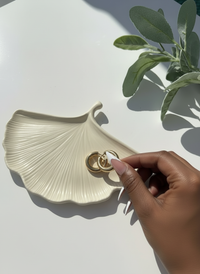 Hand holding a gold earring near the bone "Ginkgo Leaf Trinket Dish on a light surface with a plant in the corner.