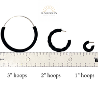 Black hoop earrings of different sizes with a ruler to scale sizing on a white background by Sunni Dineen.