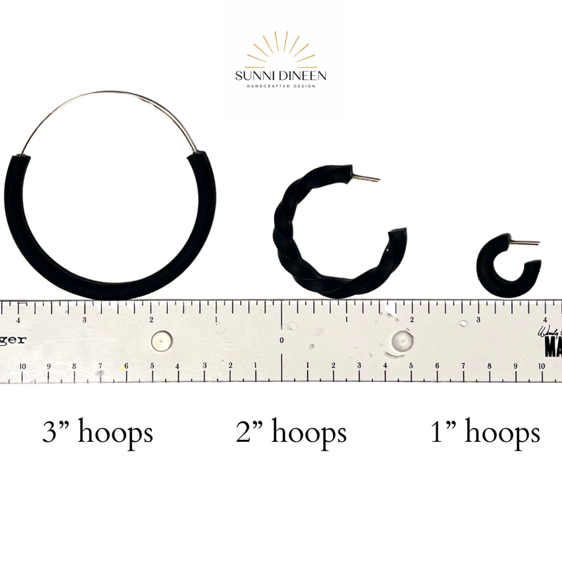 Black hoop earrings of different sizes with a ruler for scale on a white background by Sunni Dineen.