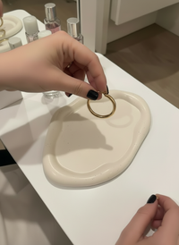Person holding a gold ring over a white cloud trinket dish handmade by Sunni Dineen, with perfume bottles in the background.