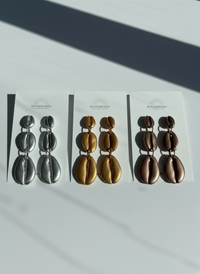 The Cowrie Shell Tiered Drop Earrings shown in silver, harvest gold, and mocha metallic displayed on Sunni Dineen Handcrafted Design earrings cards on a white surface with filtered sunlight shining down on them.