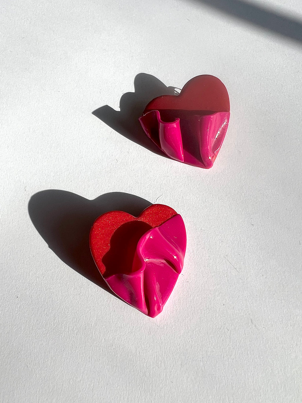 The Draped Heart Statement Earrings in red-pink displayed on a white surface, handmade by Sunni Dineen.