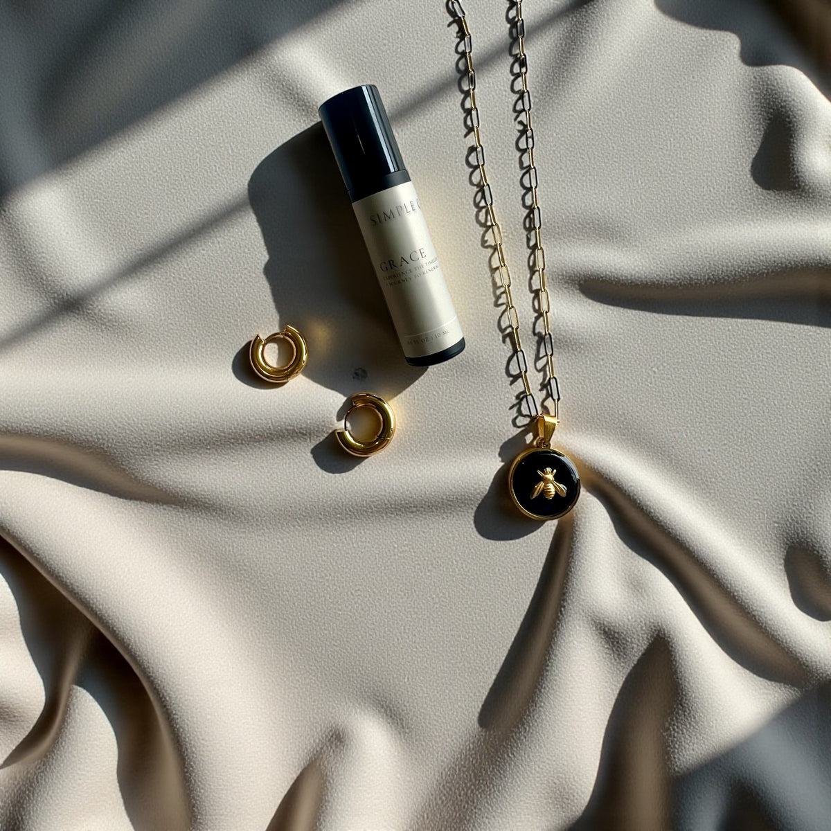 Empowered Grace Collection Set including SGB x SDHD roll-on perfume, Gold Queen Honeybee Pendant Necklace in Onyx and Chunky Gold Huggie Hoop Earrings displayed on a beige fabric background.