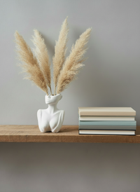 The Handcrafted Décolletage Vase made by Sunni Dineen, in bone color, displayed holding dried pampas grass on a wooden shelf with book stack.