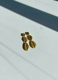 A pair of harvest gold Cowrie Shell Tiered Drop Earrings, handmade by Sunni Dineen, on a white surface with shadows.