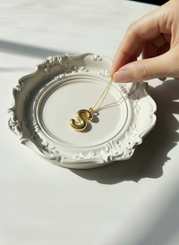 Hand holding a gold necklace with a 'S' pendant over the ivory Baroque Resin Trinket Dish handmade by Sunni Dineen.