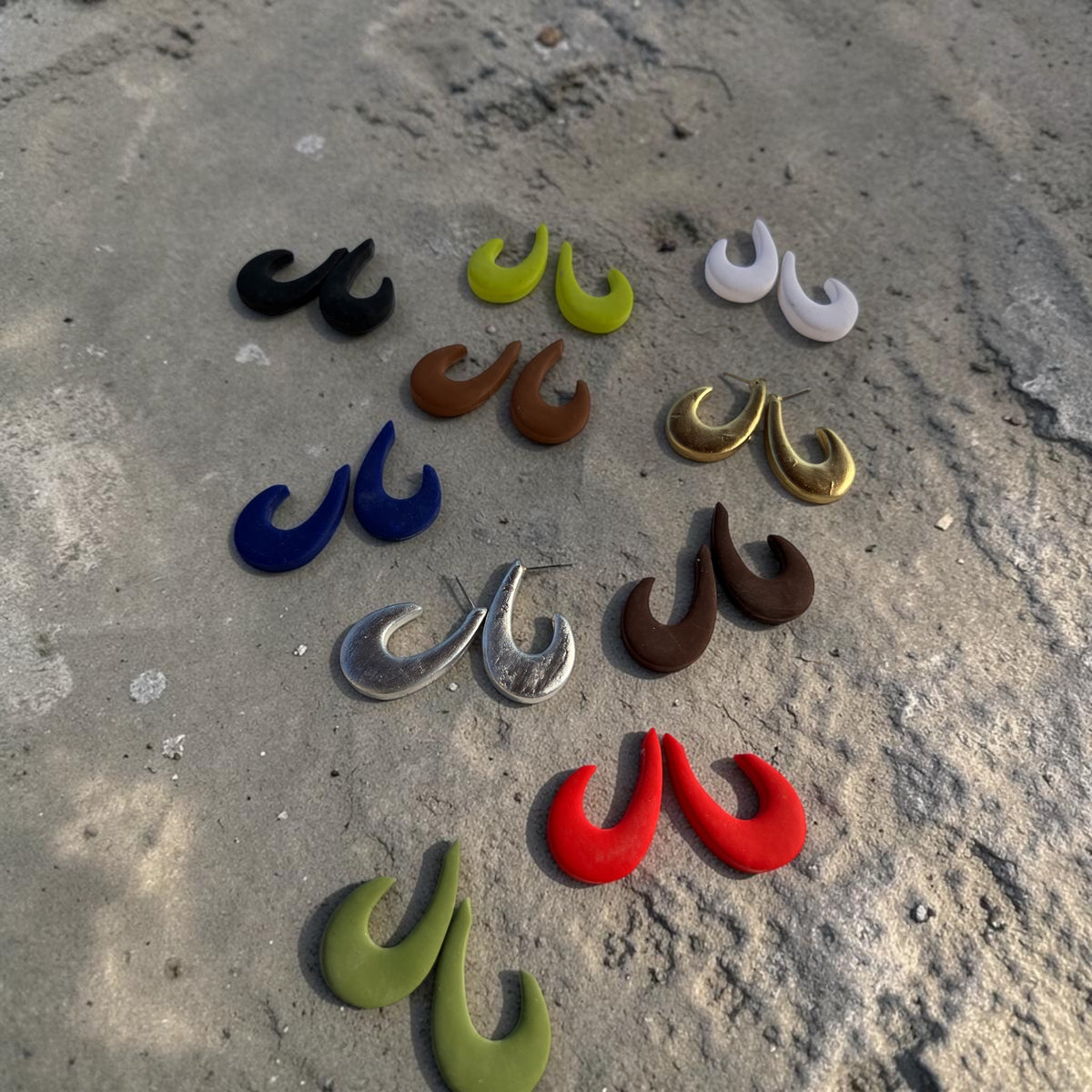 Several pairs of different colored "J Hoop Earrings" on a sandy concrete surface.