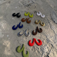 Several pairs of different colored "J Hoop Earrings" on a sandy concrete surface.
