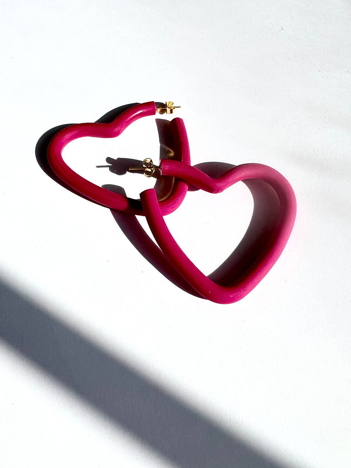 A pair of large fuchsia open heart hoop earrings on a white surface, handmade by Sunni Dineen.