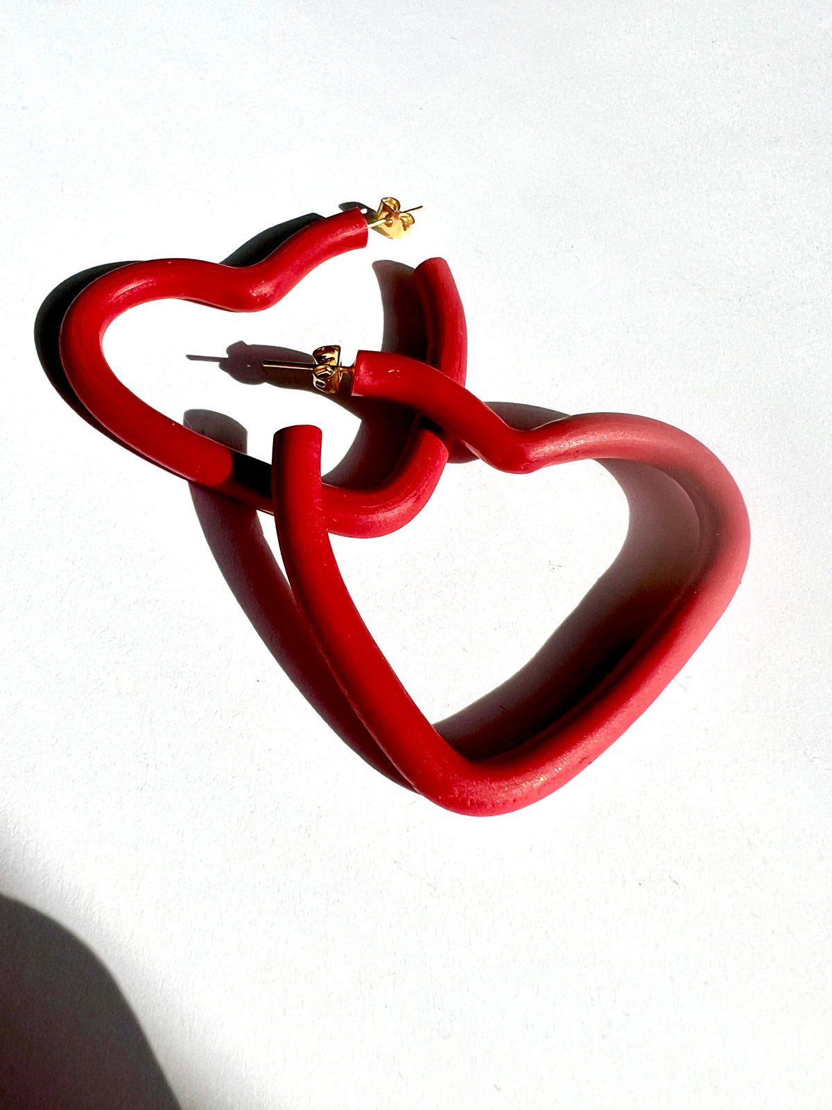A pair of large red open heart hoop earrings on a white surface, handmade by Sunni Dineen.
