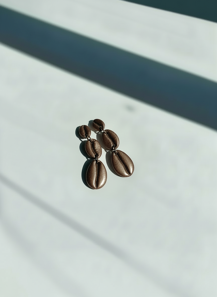 A pair of mocha metallic Cowrie Shell Tiered Drop Earrings, handmade by Sunni Dineen, on a white surface with shadows.