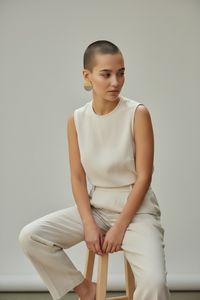 A model with a buzz cut, sitting on a wooden stool, styled wearing a white sleeveless blouse, khaki trousers, and a pair of Lumi Statement Earrrings.