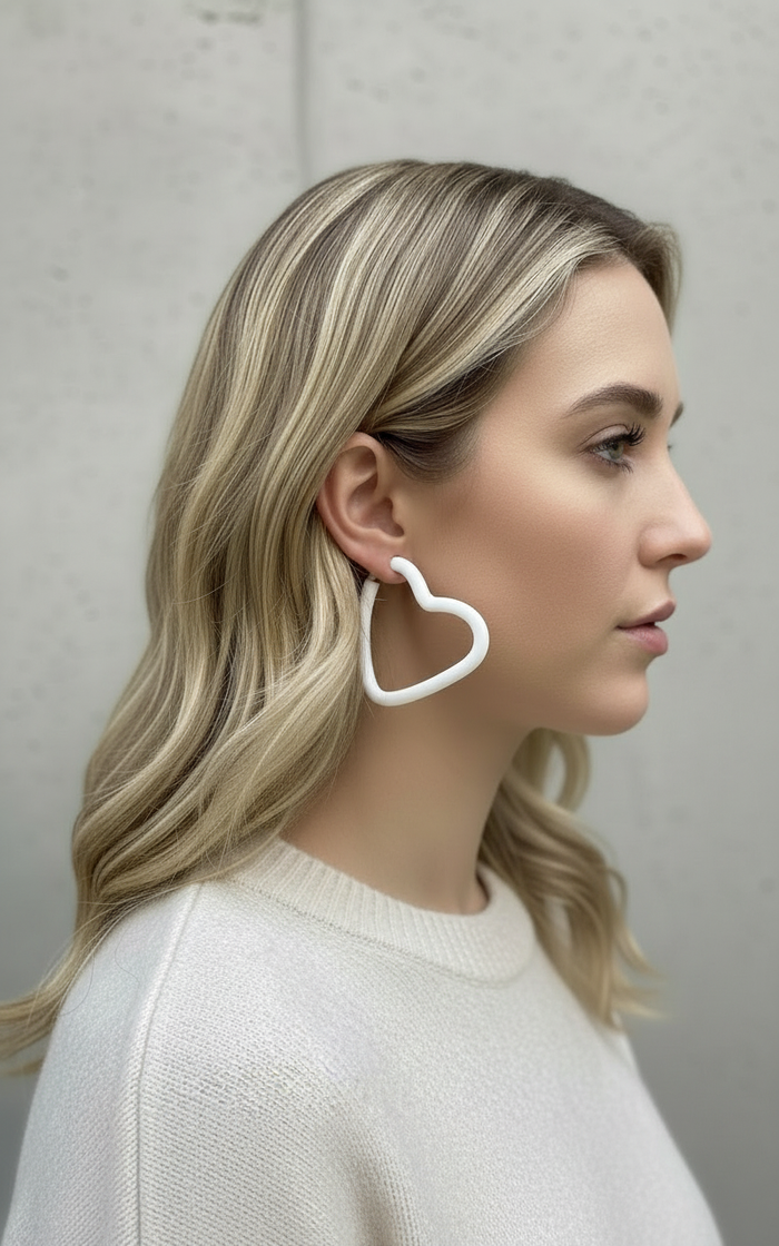 Woman wearing a pair of large white open heart hoop earrings handmade by Sunni Dineen;  against a neutral background