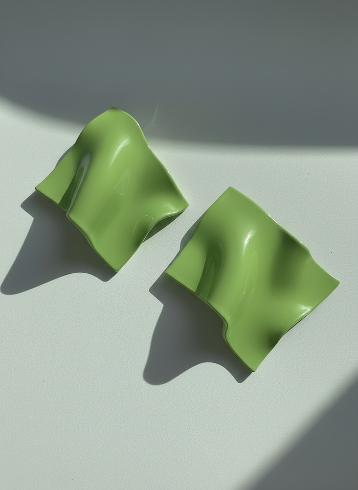 A pair of avocado 'Monae' statement earrings, handmade by Sunni Dineen, on a white surface with shadows and filtered light.