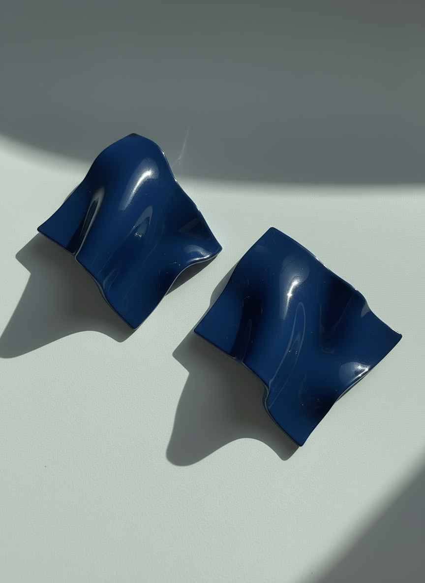 A pair of blue 'Monae' statement earrings, handmade by Sunni Dineen, on a white surface with shadows and filtered light.