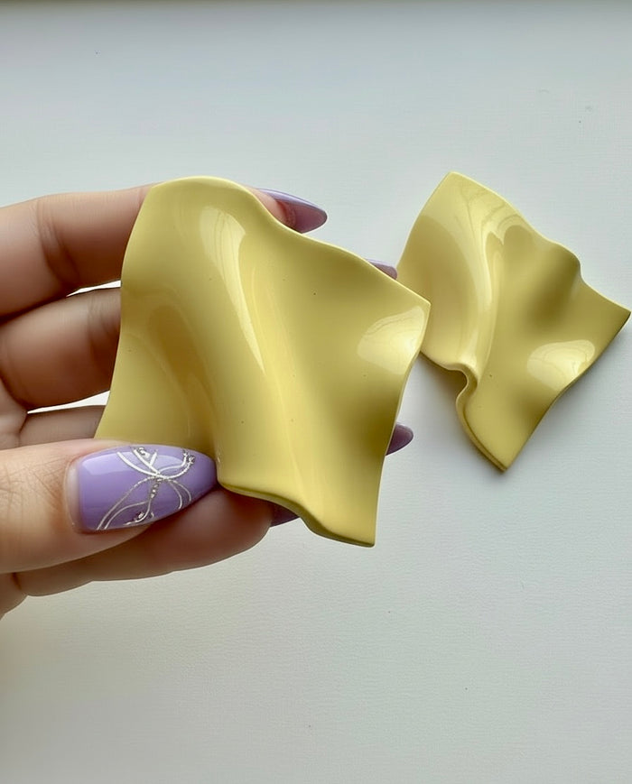 Hand holding a pair of buttercream yellow 'Monae' statement earrings, handmade by Sunni Dineen against a white surface.