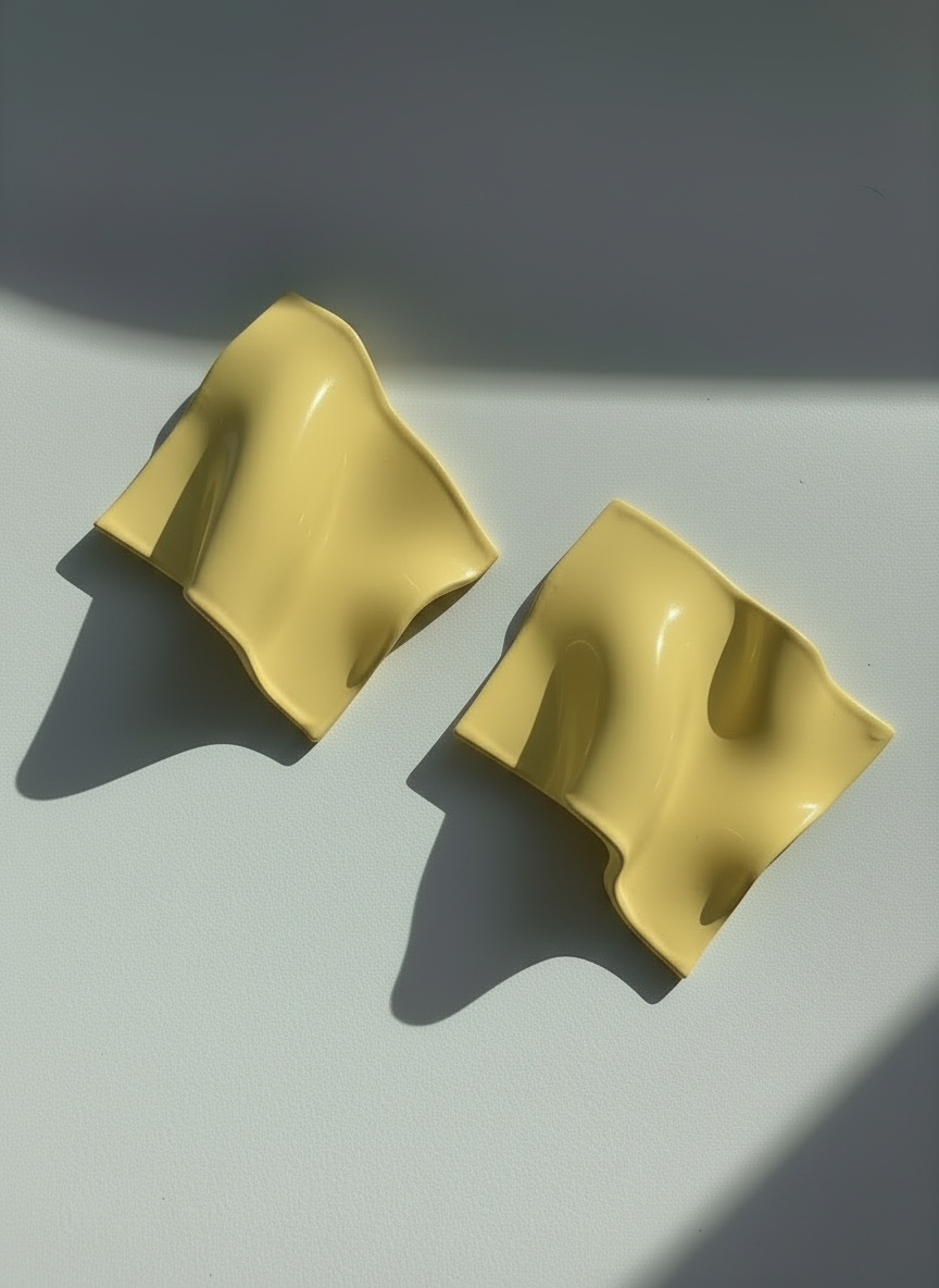 A pair of buttercream yellow 'Monae' statement earrings, handmade by Sunni Dineen, on a white surface with shadows and filtered light.