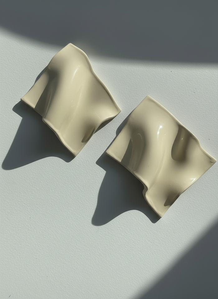 A pair of cream 'Monae' statement earrings, handmade by Sunni Dineen, on a white surface with shadows and filtered light.