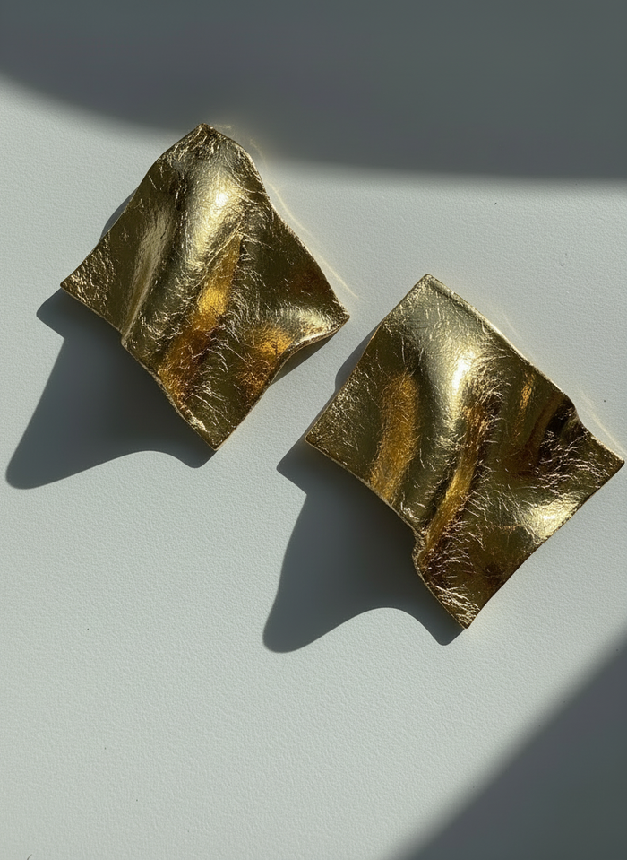 A pair of gold leaf 'Monae' statement earrings, handmade by Sunni Dineen, on a white surface with shadows and filtered light.