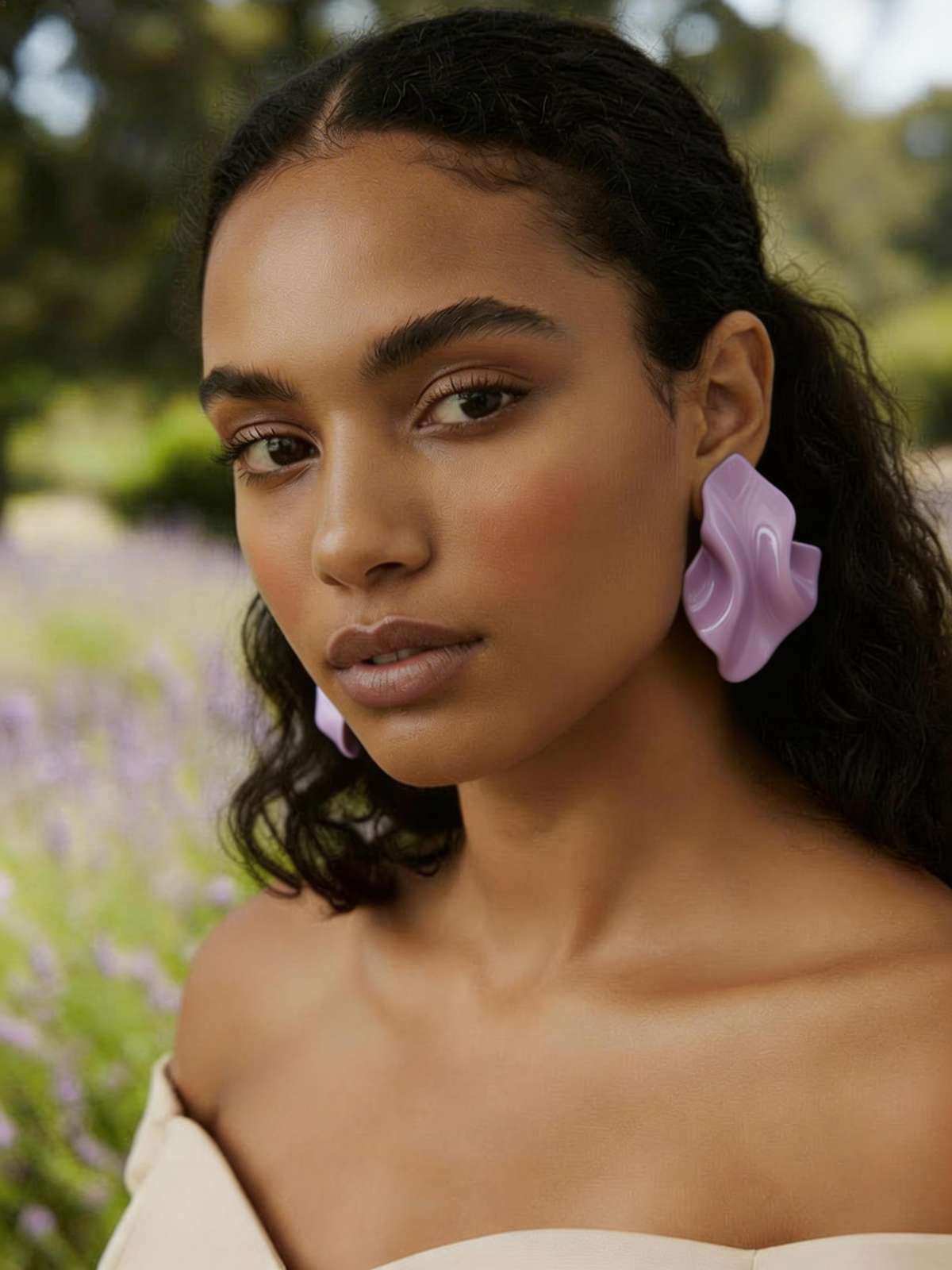 Woman wearing a pair of lavender 'Monae' statment earrings handmade by Sunni Dineen, in a natural setting with lavender flowers.