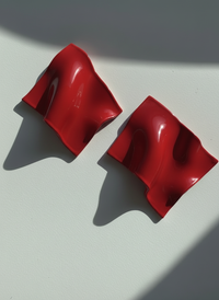 A pair of red 'Monae' statement earrings, handmade by Sunni Dineen, on a white surface with shadows and filtered light.