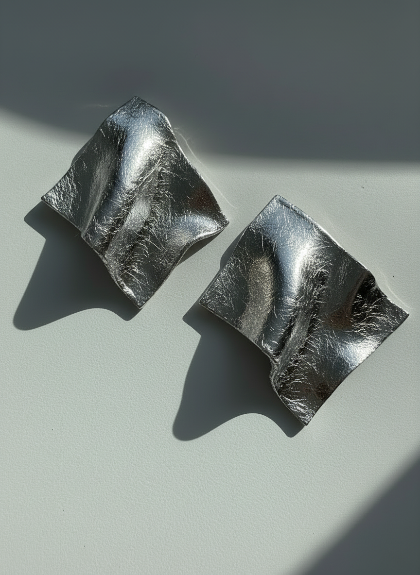 A pair of silver leaf 'Monae' statement earrings, handmade by Sunni Dineen, on a white surface with shadows and filtered light.