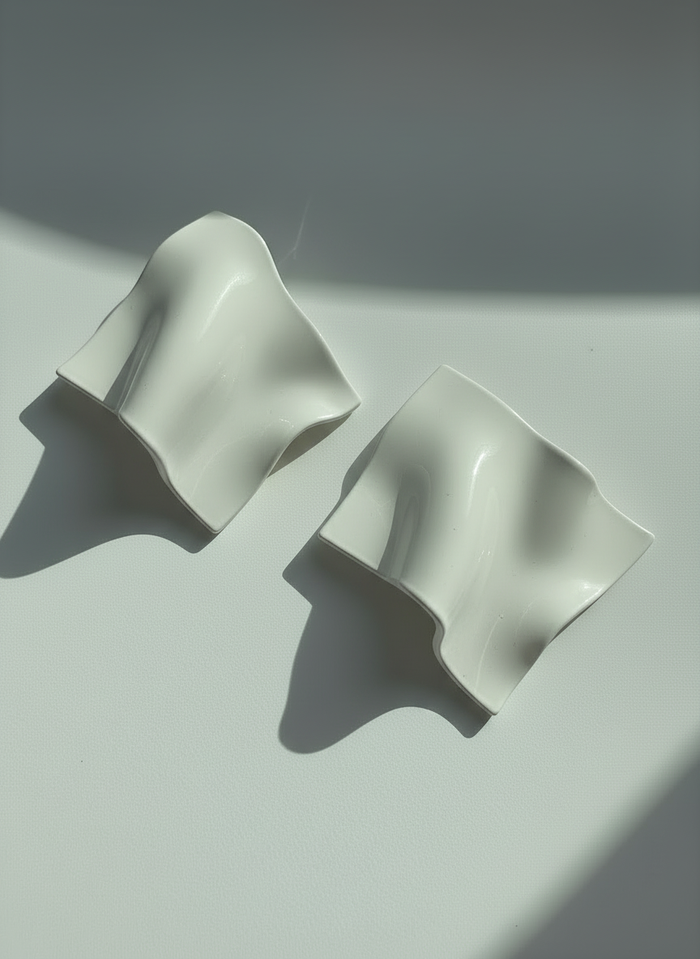 A pair of white 'Monae' statement earrings, handmade by Sunni Dineen, on a white surface with shadows and filtered light.