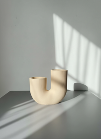 Beige U-shaped ribbed vase, handmade by Sunni Dineen on a gray surface with sunlight casting shadows on a light gray wall.