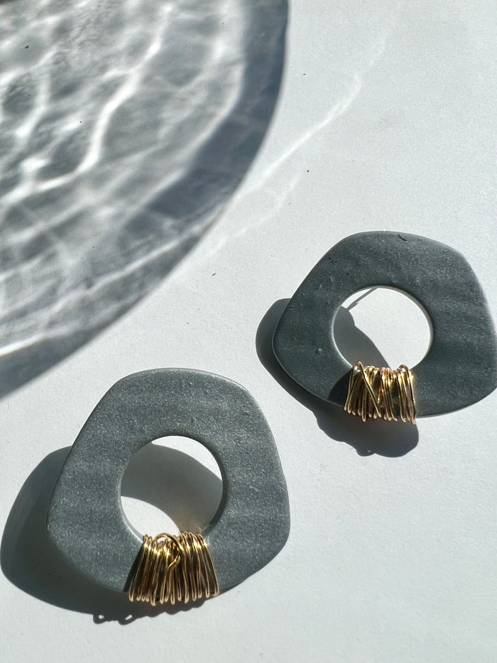 A pair of pewter-gold "Tulum" statement earrings showcased on white concrete with a pool water reflection on its surface.