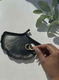 A video of a model placing a pair of gold hoop earrings on a black Ginkgo Leaf Trinket Dish.