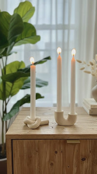 A video of the ivory Mini Sculpted Candlestick Holder Set; a knot and u-shaped candlestick holders styled with lit taper candles.