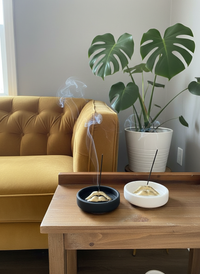 A black and white Sculptural Lips Incense Holder with incense burning on a wooden table with a yellow sofa and potted plant in the background.