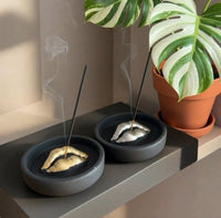 Two black Sculptural Lips Incense Holders handmade by Sunni Dineen, holding burning incense sticks, a potted plant in the background.