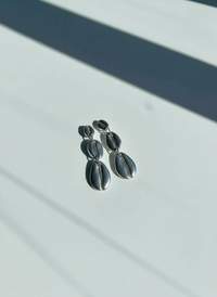 A pair of silver Cowrie Shell Tiered Drop Earrings, handmade by Sunni Dineen, on a white surface with shadows.
