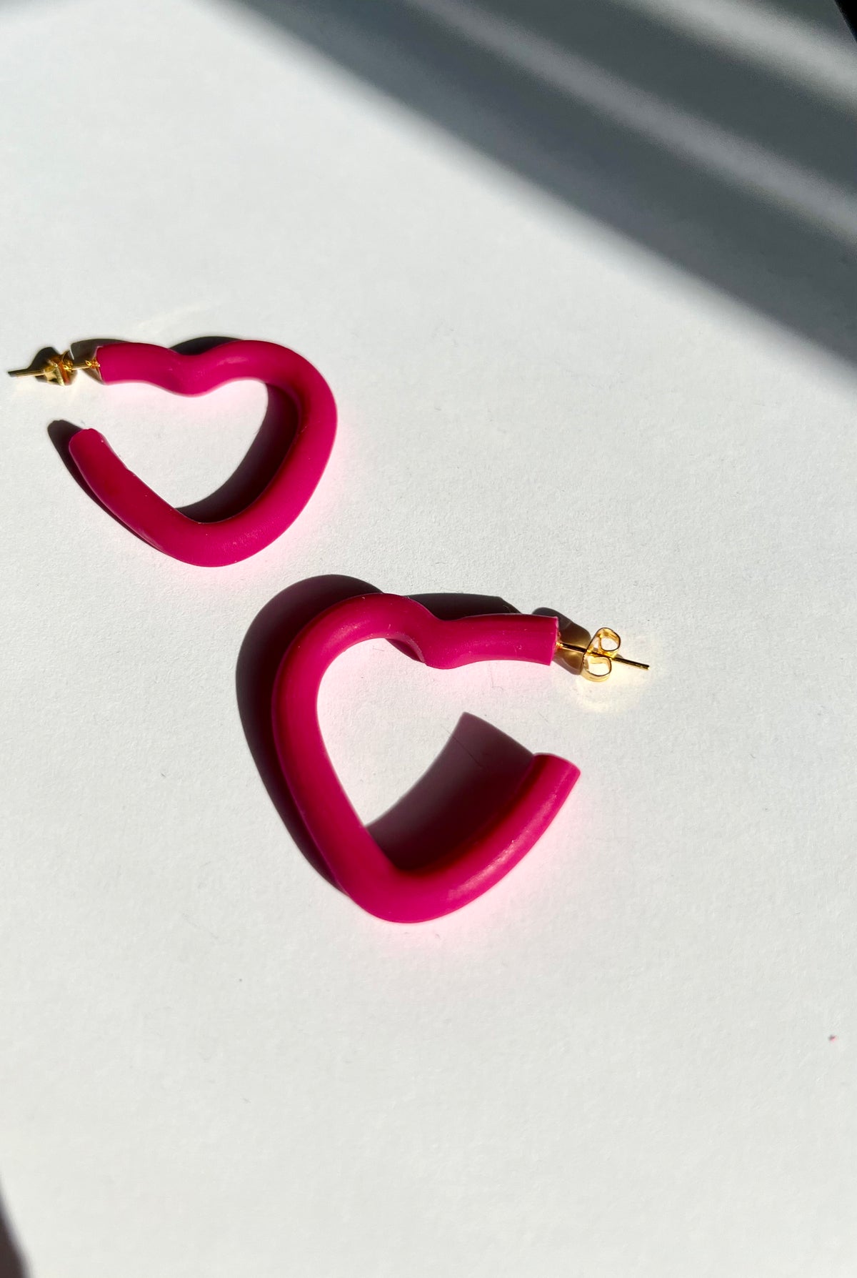 A pair of small fuchsia open heart hoop earrings on a white surface, handmade by Sunni Dineen.