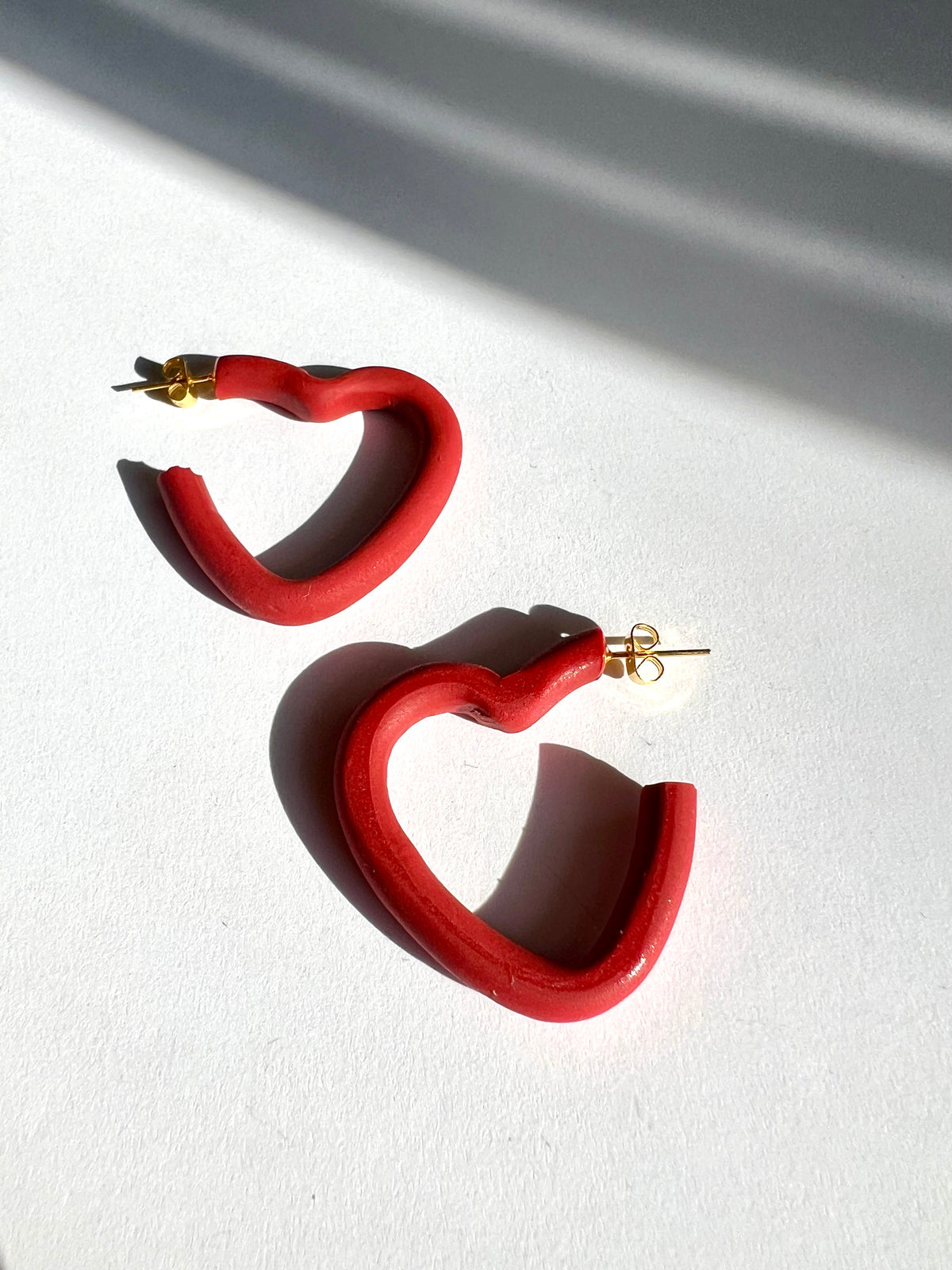 A pair of small red open heart hoop earrings on a white surface, handmade by Sunni Dineen.