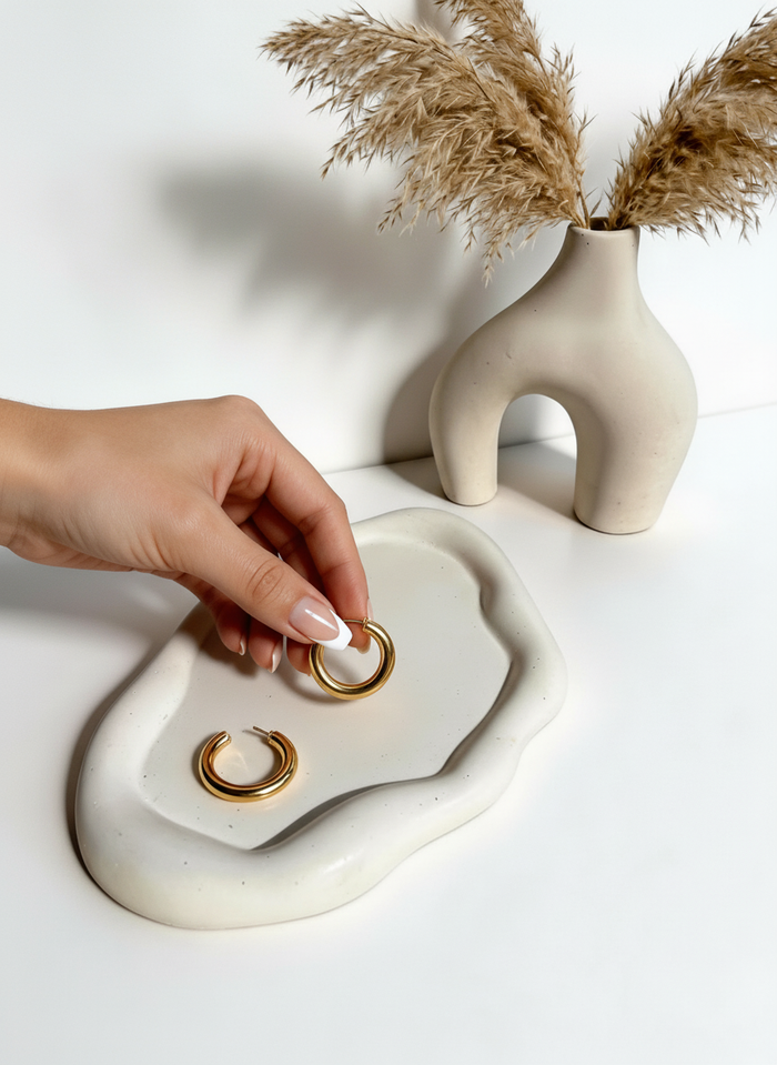 Hand holding a gold ring over the bone-colored Cloud Trinket Dish with a bone-colored Modern Sculptural Horseshoe Vase styled with dried pampas; all on a white surface.
